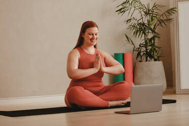 Woman doing yoga meditation watching online fitness session. Happy female in sportswear exercising at home with laptop in front. - JLPSF05753