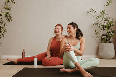 Glückliche junge Frauen, die auf dem Boden eines Fitnessstudios sitzen und lächeln. Zwei Frauen machen eine Pause vom Training im Fitnessstudio. - JLPSF05791