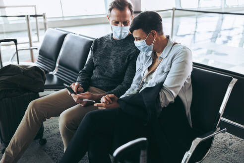 Ein Paar mit Gesichtsmasken sitzt am Flughafen und nutzt ein Mobiltelefon, um Fluginformationen abzurufen. Ein Mann zeigt einer Frau sein Mobiltelefon, während sie am Flughafen auf ihren Flug während der Pandemie wartet. - JLPSF05927