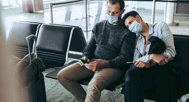 Gelangweiltes Touristenpaar, das während einer Pandemie auf dem Flughafen festsitzt. Ein Mann telefoniert mit einer Frau auf dem Flughafen, beide tragen Gesichtsmasken. - JLPSF05932