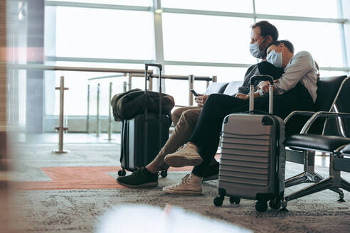 Ein Paar mit Gesichtsmasken sitzt im Flughafenterminal und wartet auf den Flug. Ein Mann benutzt ein Handy und sitzt neben einer Frau am Flughafen während der Corona-Virus-Pandemie. - JLPSF05933