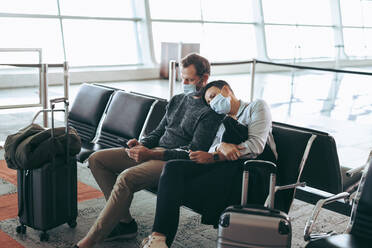 Reisendes Pärchen in Pandemie beim Warten im Flughafenterminal. Mann und Frau mit Gesichtsmasken sitzen im Wartebereich des Flughafens. - JLPSF05935