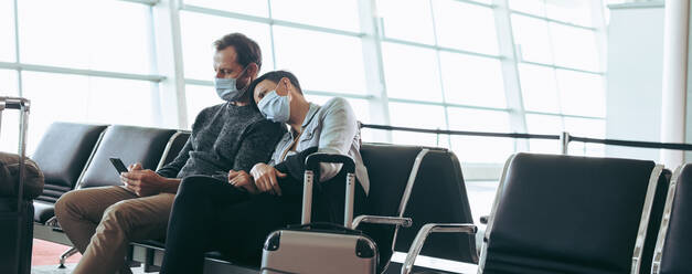 Man and woman with face masks sitting at airport terminal waiting lounge. Traveler couple stranded during corona virus outbreak at airport. - JLPSF05937