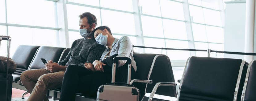 Mann und Frau mit Gesichtsmasken in der Wartelounge eines Flughafenterminals. Reisendes Paar, das während des Ausbruchs des Corona-Virus am Flughafen festsitzt. - JLPSF05937