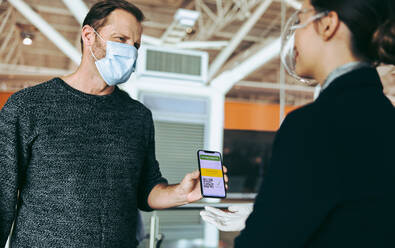 Männlicher Reisender mit Gesichtsmaske zeigt dem Bodenpersonal seinen Impfpass auf dem Smartphone. Flugbegleiter überprüft den Impfpass eines Mannes bei der Abfertigung am Flughafen während der Pandemie. - JLPSF05941