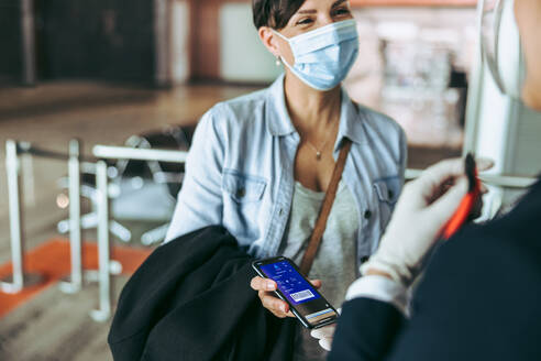 Eine Frau mit Gesichtsmaske zeigt dem Flugbegleiter ihre Bordkarte auf dem Mobiltelefon. Eine weibliche Reisende am Flughafen-Check-in-Schalter zeigt ihre Bordkarte. - JLPSF05943