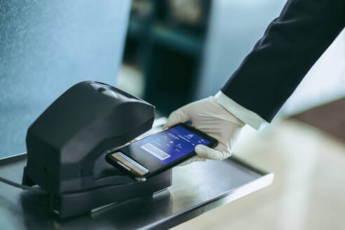 Ausgeschnittene Aufnahme einer Hand, die ihre Bordkarte mit dem Handy unter den Scanner am Flughafenschalter legt. Hand eines Flughafenmitarbeiters mit Handschuh, der die Bordkarte während einer Pandemie scannt. - JLPSF05955
