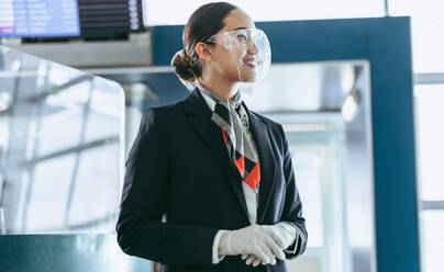 Air hostess in face shield standing at airport. Airport employee at terminal during pandemic waiting for passengers. - JLPSF05958