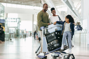 Junge Tochter sitzt mit ihren Eltern auf einem Gepäckwagen am internationalen Flughafen. Afrikanische Familie wartet am Flughafen auf ihren Flug. - JLPSF05972