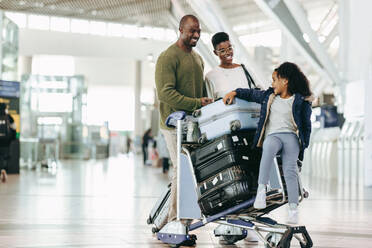 Young daughter sitting on luggage trolley with her parents at international airport. African family waiting at airport for their flight. - JLPSF05972