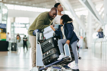 Afrikanische Familie mit Gepäckwagen, die im Flughafenterminal auf ihren Flug wartet. Der Vater unterhält sich mit seiner Tochter, die auf dem Gepäckwagen sitzt, während die Mutter die Flugzeiten am Flughafen überprüft. - JLPSF05973