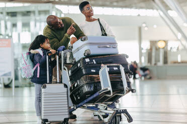 Vater zeigt seiner Tochter die Fluginformationstafel im Flughafenterminal. Junge Familie, die mit ihrem Gepäck auf einem Trolley auf den Flug wartet. - JLPSF05975