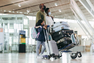 Afrikanische Familie, die im Flughafenterminal steht und auf ihren Flug wartet. Eltern mit Tochter mit Gesichtsmasken am Flughafen mit ihrem Gepäck auf einem Trolley. - JLPSF05976