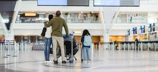 Rückansicht einer Familie mit Gepäckwagen, die im Flughafenterminal auf ihren Flug wartet. Eltern mit kleiner Tochter, die am Flughafen stehen und die Flugzeiten überprüfen. - JLPSF05977