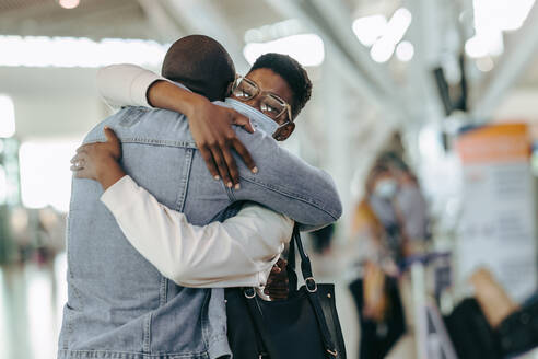 Afrikanisches Reisendenpaar, das sich am Flughafen umarmt. Mann am Flughafen-Terminal, der eine Frau umarmt, bevor er sich während einer Pandemie verabschiedet. - JLPSF05978