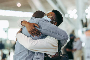 Ein Mann umarmt eine Frau während einer Pandemie im Flughafenterminal. Ein afrikanisches Paar trifft sich bei der Ankunft am Flughafen. - JLPSF05979