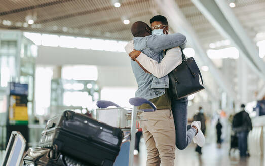 Afrikanischer Mann umarmt und hebt Frau im Flughafenterminal hoch. Paar trifft sich nach langer Trennung auf dem Flughafen nach der Pandemie-Sperre. - JLPSF05983