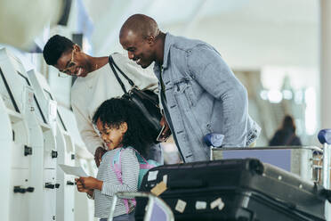 African family looking at the boarding pass in hands of their daughter at airport. Young girl looking happy to hold air ticket print while standing with mother and father at airport. - JLPSF05985
