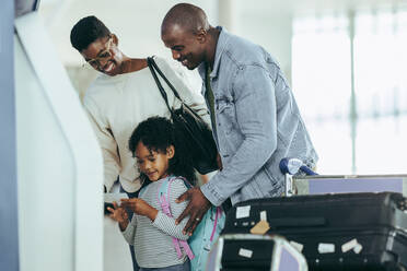 African young girl with family reading boarding pass print from machine at airport. Cute girl holding air ticket print while standing with mother and father at airport. - JLPSF05986