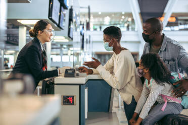 Fluggäste, die während der Pandemie mit dem Flughafenpersonal am Check-in-Schalter stehen. Afrikanische Familie bei Ausbruch von Covid-19 am Check-in-Schalter des Flughafens. - JLPSF05989