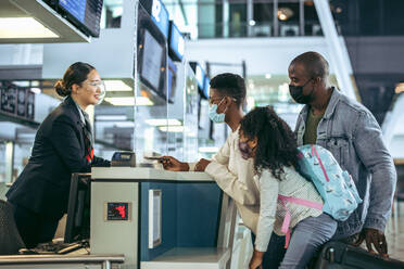 Touristen am Check-in-Schalter mit Flugbegleiter während der Pandemie. Afrikanische dreiköpfige Familie in der Pandemiezeit bei der Passübergabe am Check-in-Schalter des Flughafenterminals. - JLPSF05990