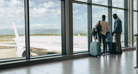 Weitwinkelansicht einer dreiköpfigen Familie, die mit ihrem Gepäck im Flughafenterminal steht und die Flugzeuge durch ein Glasfenster beobachtet. Reisende, die am Flughafenfenster mit Gepäck auf ihren Flug warten. - JLPSF05992