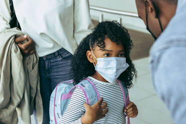 Afrikanisches Mädchen mit Gesichtsmaske und Rucksack schaut ihren Vater an. Niedliches Mädchen mit chirurgischer Gesichtsmaske am Flughafen. - JLPSF05994