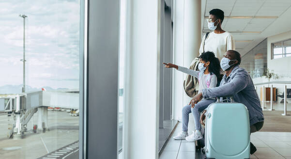 Familie mit Gepäck wartet am Flughafen-Terminal und beobachtet Flugzeuge durch ein Glasfenster. Junges Mädchen zeigt ihren Eltern ein Flugzeug am Flughafen. - JLPSF05995