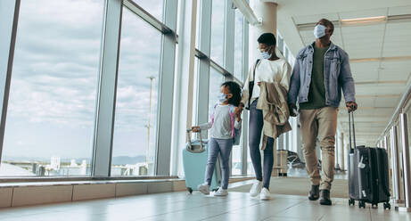 Afrikanische Familie mit Gepäck, die am Fenster des Flughafenterminals vorbeigeht und die Flugzeuge durch das Glasfenster beobachtet. Familie, die durch den Flughafengang geht. - JLPSF05996