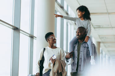 Glückliche afrikanische Familie auf dem Weg in den Urlaub im Flughafen. Ehepaar zu Fuß im Flughafen mit Tochter sitzt auf der Schulter und zeigt weg. - JLPSF06000