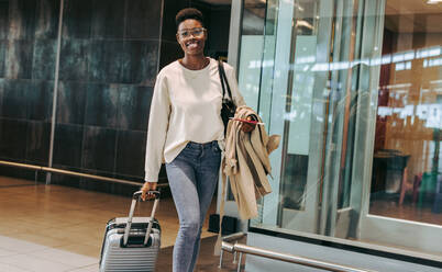 African woman walking at airport with trolley bag and coat. Female tourist carrying luggage while walking at international airport. - JLPSF06002