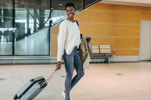 Afrikanische Frau mit Trolley und Mantel, die beim Gehen auf dem Flughafen zurückblickt. - JLPSF06003