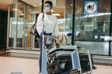 Woman in face mask pushing luggage trolley while walking at airport. Female passenger traveling in pandemic. - JLPSF06004