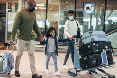 Ein Mann und eine Frau mit einem kleinen Mädchen, die während der Pandemie am Flughafen unterwegs sind. Eine dreiköpfige afrikanische Familie geht mit Gesichtsmasken am Flughafenterminal vorbei. - JLPSF06005