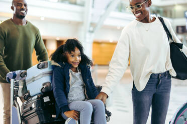 Ein Mann und eine Frau mit ihrer kleinen Tochter sitzen auf einem Gepäckwagen am Flughafen. Eine Touristenfamilie im Flughafenterminal auf dem Weg in den Urlaub. - JLPSF06010