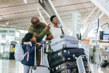 Ein Mann zeigt seiner Tochter etwas, während er mit seinem Gepäck am Flughafen steht. Eine dreiköpfige afrikanische Familie wartet im Flughafenterminal auf ein Flugzeug. - JLPSF06013