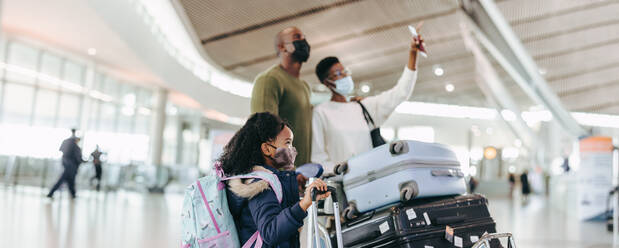 Mutter, die ihrer Familie am Flughafen etwas zeigt. Junge Tochter mit Gepäckwagen und Eltern im Hintergrund am Flughafen während einer Pandemie. - JLPSF06014