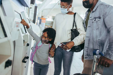 Dreiköpfige Familie am Flughafen in der Nähe des Selbstbedienungs-Check-in-Automaten während der Pandemie. Afrikanisches Paar mit süßer Tochter am Flughafen. - JLPSF06021