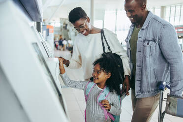 Kleines Mädchen freut sich mit ihren Eltern über den Selbstbedienungs-Check-in-Automaten am Flughafen. Dreiköpfige Familie am Check-in-Automaten eines internationalen Flughafens. - JLPSF06022