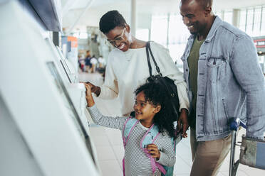 Small girl happy to use self service check in machine at airport with her parents. Family of three at international airport automated check in machine. - JLPSF06022