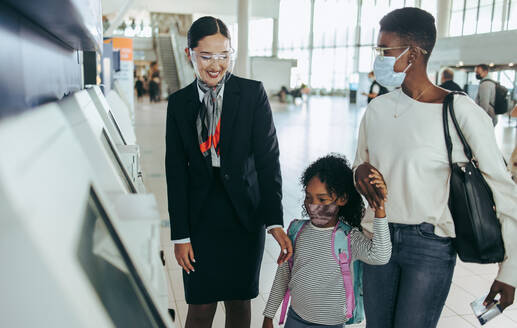 Ein Flugbegleiter hilft einem Mädchen und seiner Mutter mit dem Selbstbedienungs-Check-in-Automaten. Eine afrikanische Familie mit Gesichtsmaske am Flughafen mit Mitarbeitern der Fluggesellschaft. - JLPSF06024