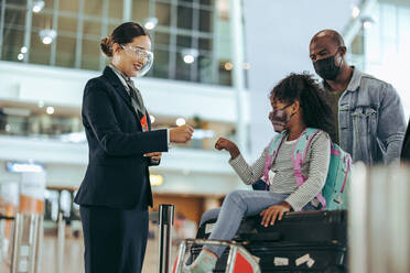Flughafenpersonal während der Pandemie, das einem jungen Mädchen aus einer Reisendenfamilie am Flugsteig die Hand gibt. Bodenpersonal am Flughafen, das ein Mädchen mit Gesichtsmaske begrüßt, das auf dem Gepäck sitzt. - JLPSF06029