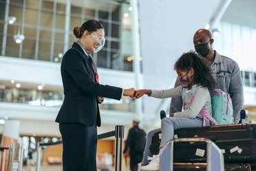Flugbegleiter und junges Mädchen aus einer Familie von Reisenden geben sich am Flugsteig die Hand. Bodenpersonal am Flughafenterminal begrüßt Mädchen mit Gesichtsmaske und ihren Vater während einer Pandemie. - JLPSF06030