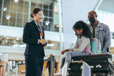 Flughafenpersonal am Flugsteig mit einem Mädchen, das auf dem Gepäck sitzt, und einem Vater, der den Gepäckwagen schiebt. Bodenpersonal am Flughafen, das einer Touristenfamilie mit Gesichtsmasken während der Pandemie hilft. - JLPSF06031