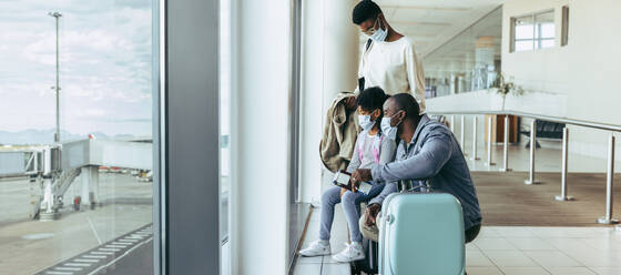 Touristenfamilie, die während der Pandemie am Fenster des Flughafens wartet. Dreiköpfige Familie am Flughafen, die vor dem Abflug des Flugzeugs Zeit miteinander verbringt. - JLPSF06033