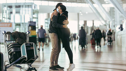 Eine Frau mit Gesichtsmaske empfängt einen Mann bei der Ankunft am Flughafen. Ein Paar trifft sich und umarmt sich am Ankunftsgate des Flughafens. - JLPSF06048