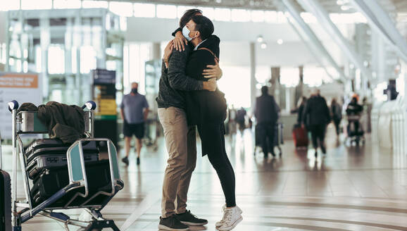 Eine Frau mit Gesichtsmaske empfängt einen Mann bei der Ankunft am Flughafen. Ein Paar trifft sich und umarmt sich am Ankunftsgate des Flughafens. - JLPSF06048