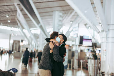 Reisender Mann umarmt Frau am Ankunftsgate des Flughafens. Frau mit Gesichtsmaske begrüßt Mann nach der Ankunft von einer Pandemie-Tour. - JLPSF06055