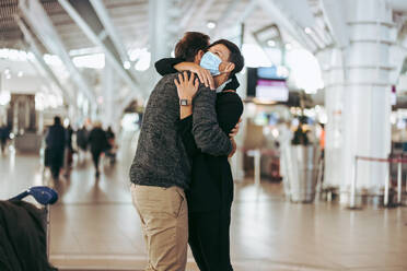 Woman in face mask giving welcome hug to man at airport arrival gate. Woman wearing face mask welcoming and embracing her boyfriend at airport - JLPSF06056