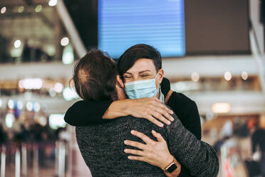 Ehemann und Ehefrau umarmen sich auf dem Flughafen während der Pandemie. Frau mit Gesichtsmaske, die ihren Mann auf dem Flughafen während der Pandemie begrüßt. - JLPSF06058